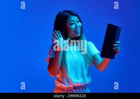Femme asiatique souriante agitant bonjour tout en tenant une réunion vidéo dans la tablette numérique sur fond bleu dégradé dans la lumière néon. Banque D'Images