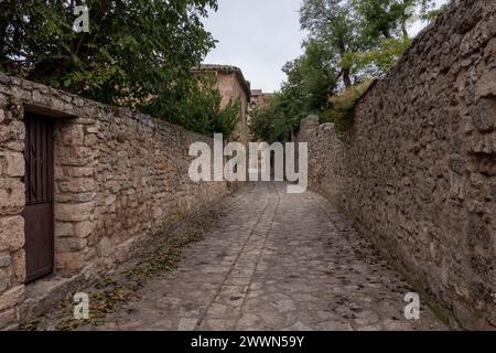 rue étroite pavée de pierre flanquée de vieux murs de pierre texturés, menant à des bâtiments anciens, sous un ciel couvert Banque D'Images