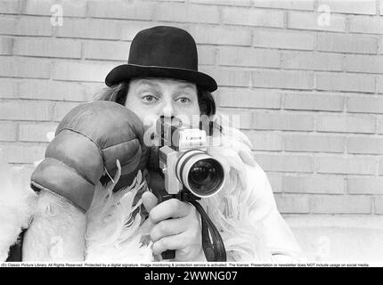 Dans les années 1970 Un homme filme avec une caméra amateur. Le film a ensuite été développé et montré sur un projecteur de film sur un écran pliant à la maison. Ricky Bruch (1946-2011) athlète suédois. Il était connu pour être un peu un showman représenté ici portant des gants de boxe, un boa en plumes et un chapeau. 1975 Banque D'Images