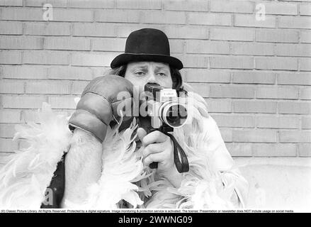Dans les années 1970 Un homme filme avec une caméra amateur. Le film a ensuite été développé et montré sur un projecteur de film sur un écran pliant à la maison. Ricky Bruch (1946-2011) athlète suédois. Il était connu pour être un peu un showman représenté ici portant des gants de boxe, un boa en plumes et un chapeau. 1975 Banque D'Images
