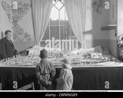 Trains miniatures dans les années 1950 Un homme à son chemin de fer miniature monté sur une table dans sa chambre. Il manipule les trains, à quelle vitesse il va et s'il veut que le train avance ou recule. Le chemin de fer modèle a des maisons, des arbres, des animaux et des gens en elle comme modèles réduits. Les enfants sont vus debout sur le côté en train de le regarder. 1952 Banque D'Images