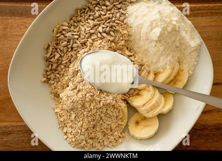 Gros plan d'une assiette profonde avec des ingrédients de muesli, comme de l'avoine, des graines de tournesol, des tranches de banane, de la poudre de protéines et une cuillerée de yaourt. La table se tient debout Banque D'Images