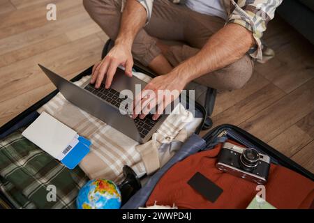 Gros plan de l'homme, mains masculines utilisant un ordinateur portable sur le dessus de la valise ouverte, emballage pour des vacances, partir en vacances, préparer l'appareil photo, deux billets d'avion Banque D'Images