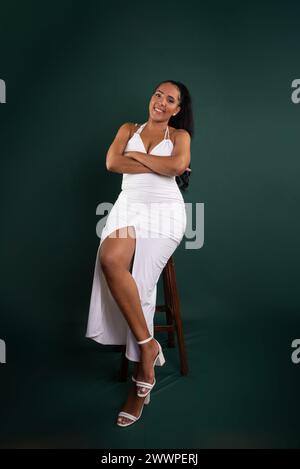 Belle femme brune regardant la caméra assise sur un tabouret en bois avec les bras croisés. Isolé sur fond vert. Banque D'Images