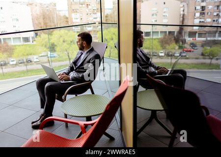 Un homme concentré en costume utilise son ordinateur portable sur un balcon ensoleillé donnant sur le paysage urbain Banque D'Images