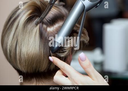 Coiffeur fait coiffure fille avec cheveux bruns dans un salon de beauté. Créez des boucles avec des fers à friser. Soins capillaires professionnels. Banque D'Images