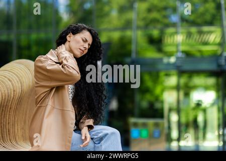 Vue latérale de la femme fatiguée souffrant de douleur aiguë dans le cou tout en ayant le temps de repos sur le banc en bois près du bâtiment vitreux. Femme bouleversée avec les yeux fermés massant la zone mal à l'extérieur. Banque D'Images