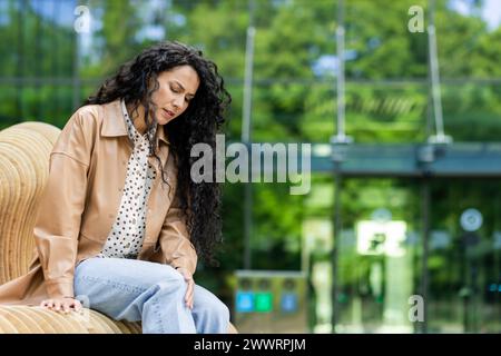 Femme caucasienne fatiguée avec de longs cheveux bouclés noirs assis sur le banc à l'extérieur touchant et éprouvant un inconfort dans le genou. Arrière-plan d'une partie de la structure en verre ou du bâtiment. Banque D'Images