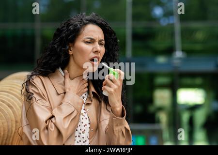 Femelle bouleversée tenant une petite bouteille verte et pulvérisant le spray de gorge dans la bouche pour soulager la douleur à l'extérieur. Dame portant manteau brun clair et chemise avec des taches brunes sur fond de bâtiment vitreux. Banque D'Images