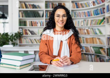 Tuteur en ligne contemporain assis à table dans la bibliothèque entourée d'étagères remplies de livres. Jolie femme portant une veste orange avec un sweat à capuche blanc tenant un stylo jaune sur le lieu de travail personnel. Banque D'Images