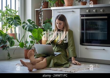 Focalisée femme intéressée freelance assise sur le sol travaillant sur ordinateur portable sur les genoux à la maison avec des plantes. Banque D'Images