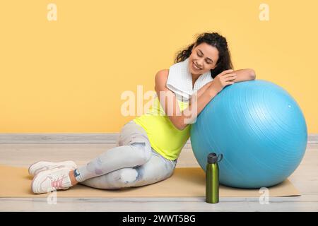 Belle jeune femme afro-américaine enceinte avec fitball et bouteille de sport sur tapis de yoga près du mur jaune Banque D'Images