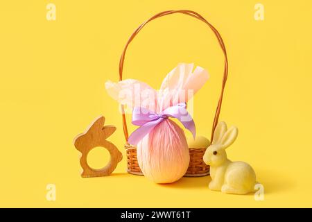 Panier en osier avec des œufs de Pâques peints et chocolat enveloppés dans du papier sur fond jaune Banque D'Images