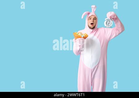 Jeune homme choqué en costume de lapin tenant un panier en osier avec des œufs de Pâques et un réveil sur fond bleu Banque D'Images