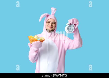 Jeune homme en costume de lapin tenant un panier en osier avec des œufs de Pâques et un réveil sur fond bleu Banque D'Images