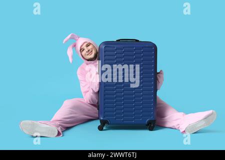 Jeune homme en costume de lapin avec valise assise sur fond bleu. Fête de Pâques Banque D'Images