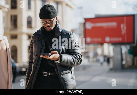 Concentré jeune entrepreneur dans les vêtements d'hiver à la mode s'engageant avec le téléphone mobile avec des panneaux d'affichage de publicité dans le fond de la ville floue. Banque D'Images