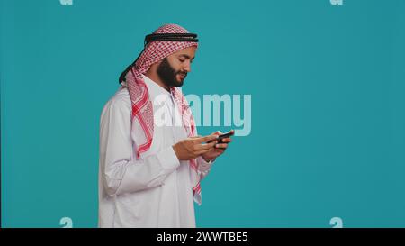 L'homme musulman porte des vêtements traditionnels et joue à des jeux vidéo sur smartphone, debout sur fond bleu. Personne arabe appréciant la concurrence de jeu mobile sur l'application de téléphone, divertissement. Banque D'Images