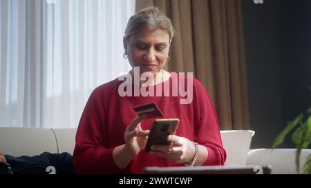 Femme d'âge moyen assis sur le canapé tenant la carte de crédit et smartphone entre le numéro de carte de crédit pour les achats en ligne. Femme utilisant instantanément facile paymen mobile Banque D'Images