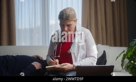 Médecin féminin senior en manteau blanc avec stéthoscope examinant le patient âgé à la maison, prescrivant des médicaments Banque D'Images