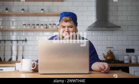 Femme en surpoids souriante entrer le numéro de carte de crédit sur ordinateur portable pour les achats en ligne, rend le paiement électronique distant facile sécurisé tout en étant assis dans la cuisine Banque D'Images