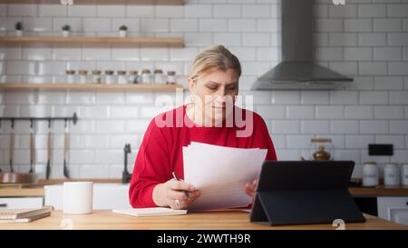 Femme d'affaires senior assis dans la cuisine à l'aide d'un appareil de tablette, vérifier les informations sur le document, prendre des notes, analyser les données, regarder à travers les papiers occupés dans PA Banque D'Images