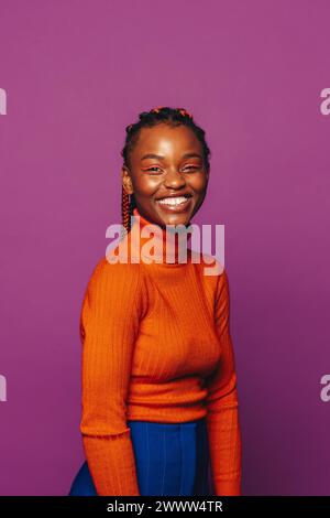 Femme heureuse et élégante avec des tresses bicolores vibrantes se tient sur un fond violet. Elle porte des vêtements décontractés, y compris un Jean et un col polo, exsud Banque D'Images