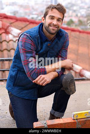 Maçon, sourire et construction avec homme constructeur sur le toit, employé travaillant dans l'industrie industrielle et carrière d'entrepreneur. Homme confiant, heureux Banque D'Images