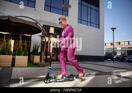 Élégamment habillée femme mature dans un pantalon rose vif et une veste roule autour de la ville sur un scooter, un frien actif, sain et éco Banque D'Images