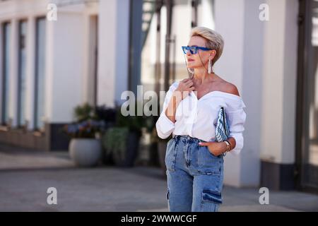 Belle femme mature de mode blonde à l'extérieur dans la rue de la ville portant chemisier blanc à la mode, pantalons de jeans cargo et accessoires à la mode, stylets Banque D'Images