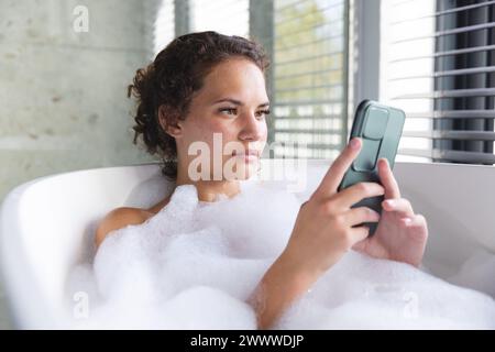 Une jeune femme biraciale se détend dans un bain moussant à la maison, en utilisant son smartphone Banque D'Images