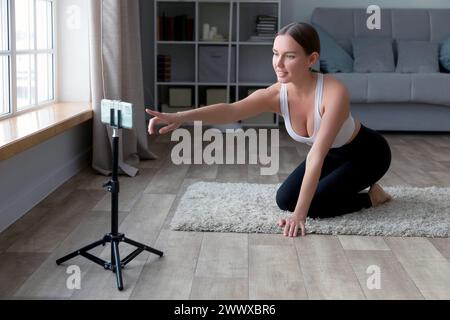 femme assise sur le sol dans un pantalon de yoga enregistrant une vidéo de fitness en ligne avec son téléphone et son trépied, souriant à la caméra dans un cadre de salon Banque D'Images