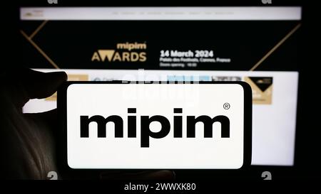 Personne tenant le téléphone portable avec le logo de la conférence immobilière française MIPIM devant la page Web de l'entreprise. Concentrez-vous sur l'affichage du téléphone. Banque D'Images