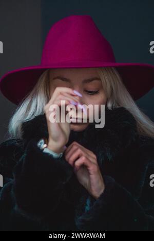 Femme blonde platine caucasienne avec des ongles curés dans un chapeau rouge regardant vers le bas réfléchi, illuminé par un éclairage de humeur changeante Banque D'Images