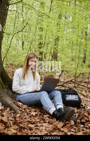 Jeune femme randonneuse avec un ordinateur portable sur ses jambes portant un pull, un Jean et des bottes de randonnée assis dans la forêt Banque D'Images