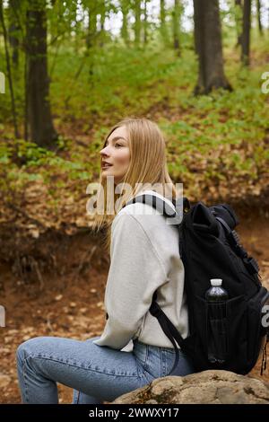 Vue de côté de confortable femme blonde souriante randonneur reposant sur les rochers, regardant la paisible vue sur la forêt Banque D'Images