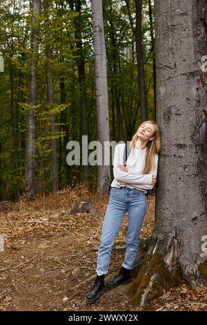 Portrait pleine hauteur de femme blonde en tenue de randonnée dans la forêt penchée sur l'arbre avec les yeux fermés Banque D'Images