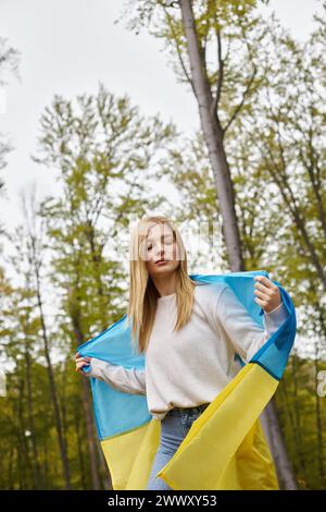 Femme blonde calme à l'extérieur dans la forêt couvrant le corps avec le drapeau national ukrainien comme cap Banque D'Images