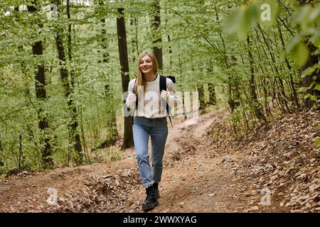 Femme blonde de randonnée heureuse portant un pull et un sac à dos marchant dans un paysage forestier dans les bois Banque D'Images