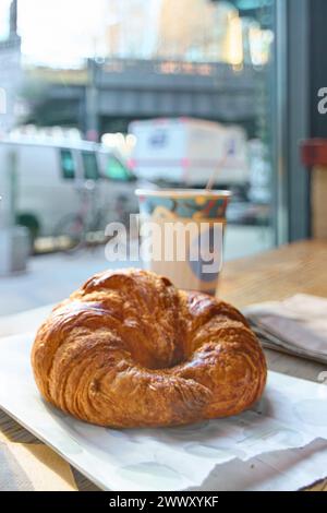 Un croissant croustillant et doré est assis sur du papier blanc sur une table en bois, avec un verre de café flou en arrière-plan, éclairé par la lumière du soleil. Banque D'Images