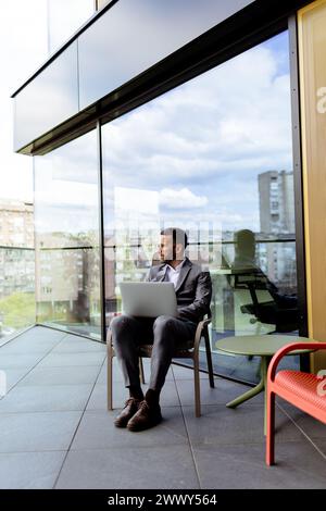 Un homme concentré en costume utilise son ordinateur portable sur un balcon ensoleillé donnant sur le paysage urbain. Banque D'Images