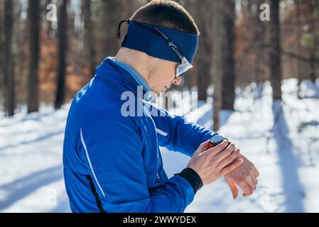 Coureur vérifiant Smartwatch pendant l'entraînement hivernal dans la forêt enneigée. Un coureur vêtu d'une veste bleue et d'un serre-tête s'arrête pour vérifier sa smartwatch lors d'une séance d'entraînement hivernale dans un cadre de forêt enneigée. Banque D'Images