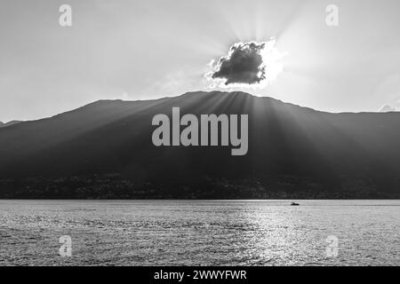 Une scène tranquille capturant la beauté sereine d'un lac de montagne au coucher du soleil. Les rayons du soleil traversent un nuage, projetant des faisceaux lumineux et des reflets étincelants sur la surface de l'eau. Un bateau solitaire profite de l'atmosphère paisible, entouré par la silhouette des montagnes. Image en noir et blanc. Banque D'Images