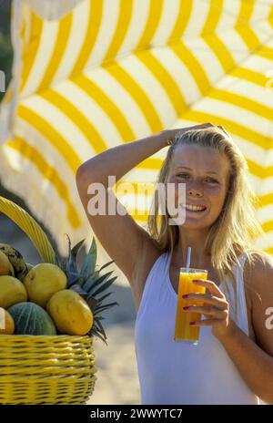 joli cheveux blonds jeune femme attrayante regardant la caméra avant heathy sourire buvant du jus d'orange un parapluie jaune panier de fruits côté sur le fond Banque D'Images