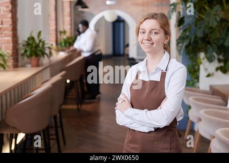 Portrait moyen d'une jeune serveuse caucasienne confiante portant une chemise blanche et un tablier brun souriant à la caméra, espace de copie Banque D'Images