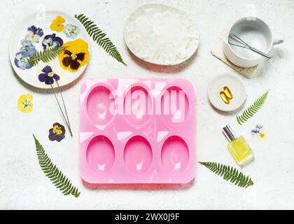 Vue de dessus du lieu de travail artisanal avec moule en silicone, fleurs pressées, feuilles de fougères, dés à base de savon et outils pour créer un savon. Décoration maison fait main con Banque D'Images