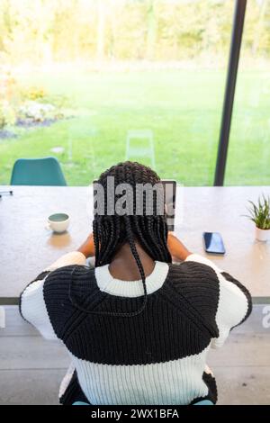 Une scène sereine avec la vue arrière d'une femme noire aux cheveux tressés travaillant attentivement sur sa tablette dans un bureau à domicile lumineux et aéré. L'espace donne sur un jardin luxuriant, alliant productivité et tranquillité de la nature. Vue arrière de la femme travaillant sur la tablette dans le bureau à domicile lumineux. Photo de haute qualité Banque D'Images