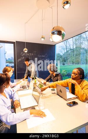 Une réunion d'équipe dynamique dans un espace de travail moderne bien éclairé. La photographie capture un groupe de professionnels ciblés, de divers horizons, en train de brainstorming autour d'une table de conférence élégante, avec un tableau noir affichant les concepts de croissance et de démarrage en arrière-plan. Équipe diversifiée engagée dans un espace de travail collaboratif. Photo de haute qualité Banque D'Images