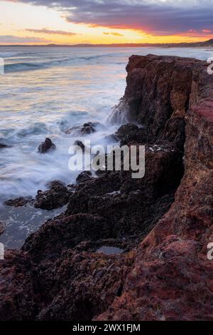 Des vagues s'écrasent sur la base d'une falaise côtière devant un ciel de coucher de soleil à Forrest Caves sur Phillip Island dans le Victoria, Australie. Banque D'Images
