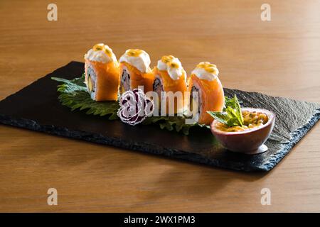 Rouleau de sushi au saumon garni de sauce crémeuse et de fruits de la passion, élégamment présenté sur une planche d'ardoise avec feuille de shiso et chou frisé ornemental - Une fusion o Banque D'Images